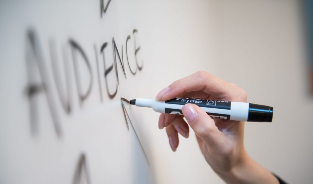 Woman's hand writing the word "audience" on a whiteboard, with arrows.
